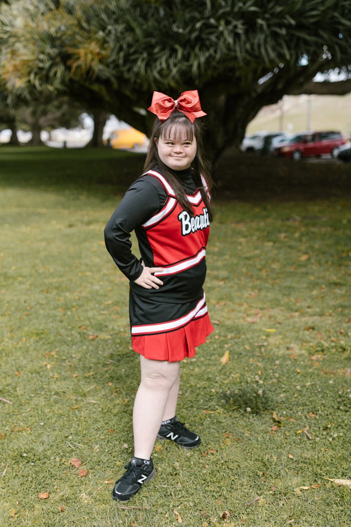 A cheerful young girl with Down Syndrome in a cheerleader outfit, posing outdoors in a park.